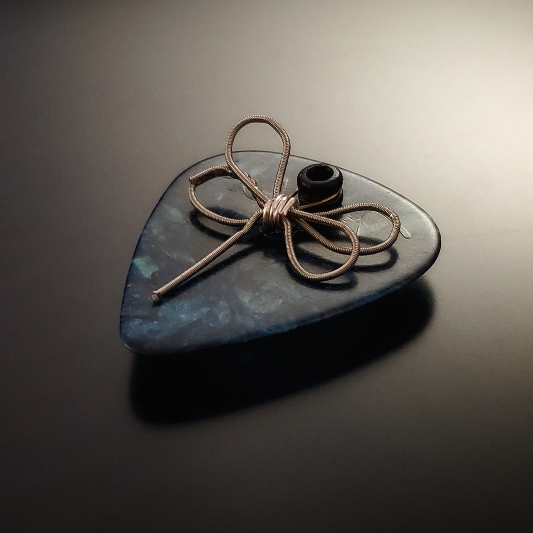 an upcycled guitar string is shaped into a dragon fly and sits on a teal guitar pick - the whole creation is a  refrigerator magnet - black background