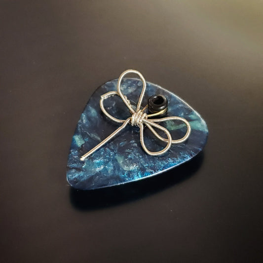 an upcycled guitar string is shaped into a dragon fly and sits on a teal guitar pick - the whole creation is a  refrigerator magnet - black background