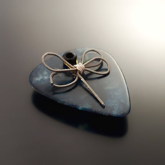 an upcycled guitar string is shaped into a dragon fly and sits on a teal guitar pick - the whole creation is a  refrigerator magnet - black background
