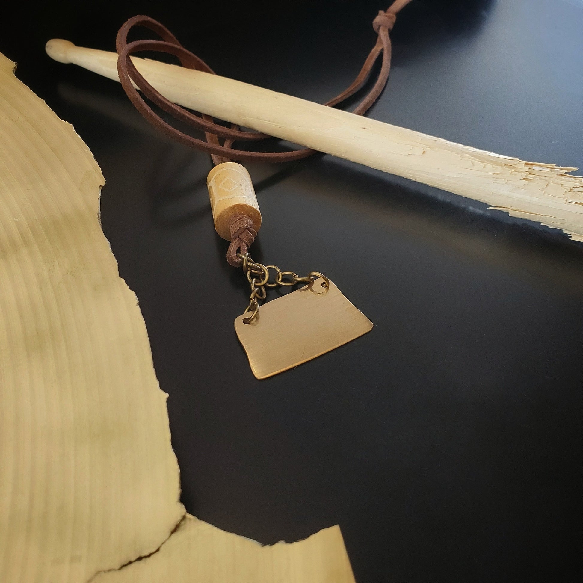 a necklace made from a piece of cymbal, chain, a piece of carved drum stick and a swede cord sits across a used drumstick, on the left there is a broken cymbal