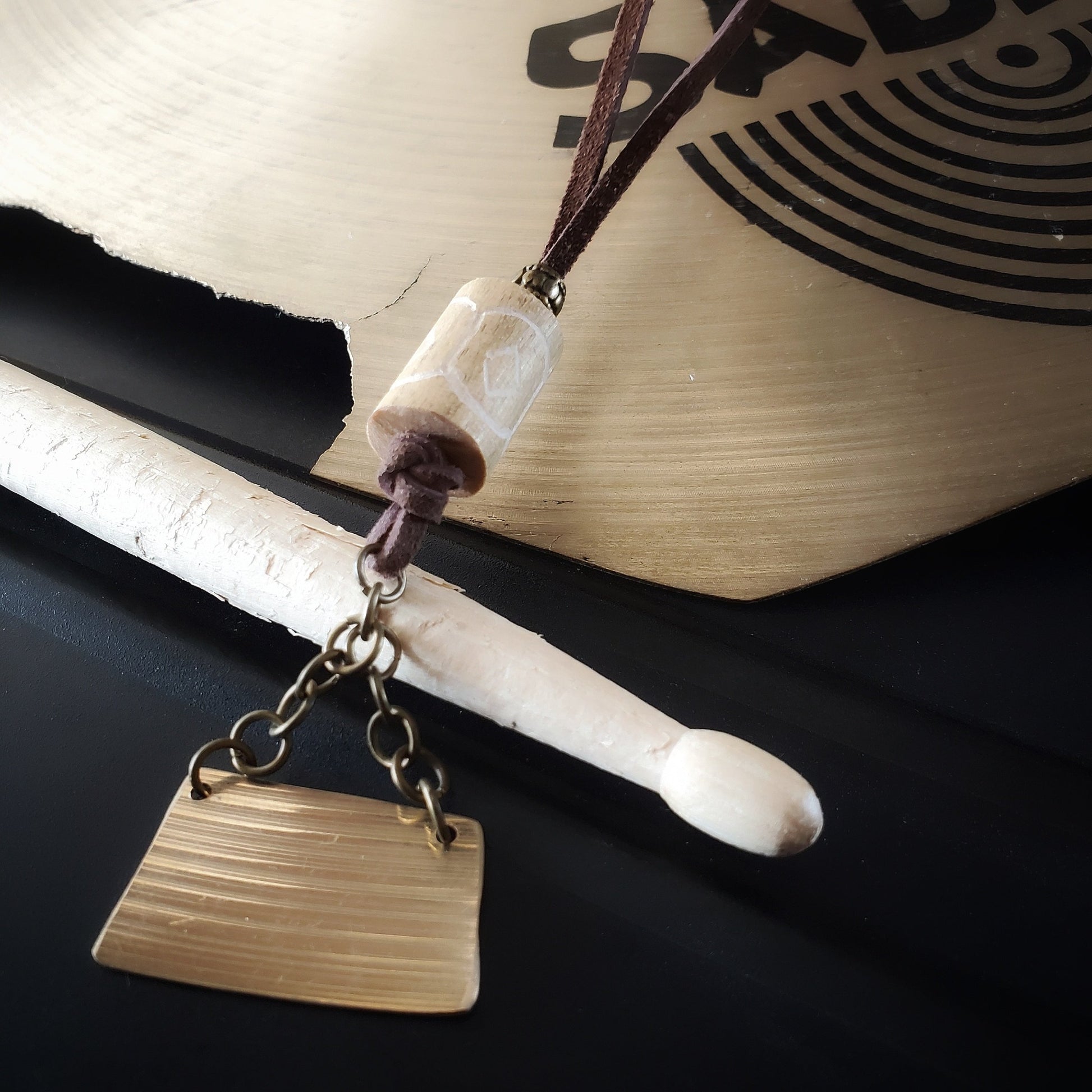 a necklace made from a piece of cymbal, chain, a piece of carved drum stick and a swede cord sits across a used drumstick and a broken cymbal