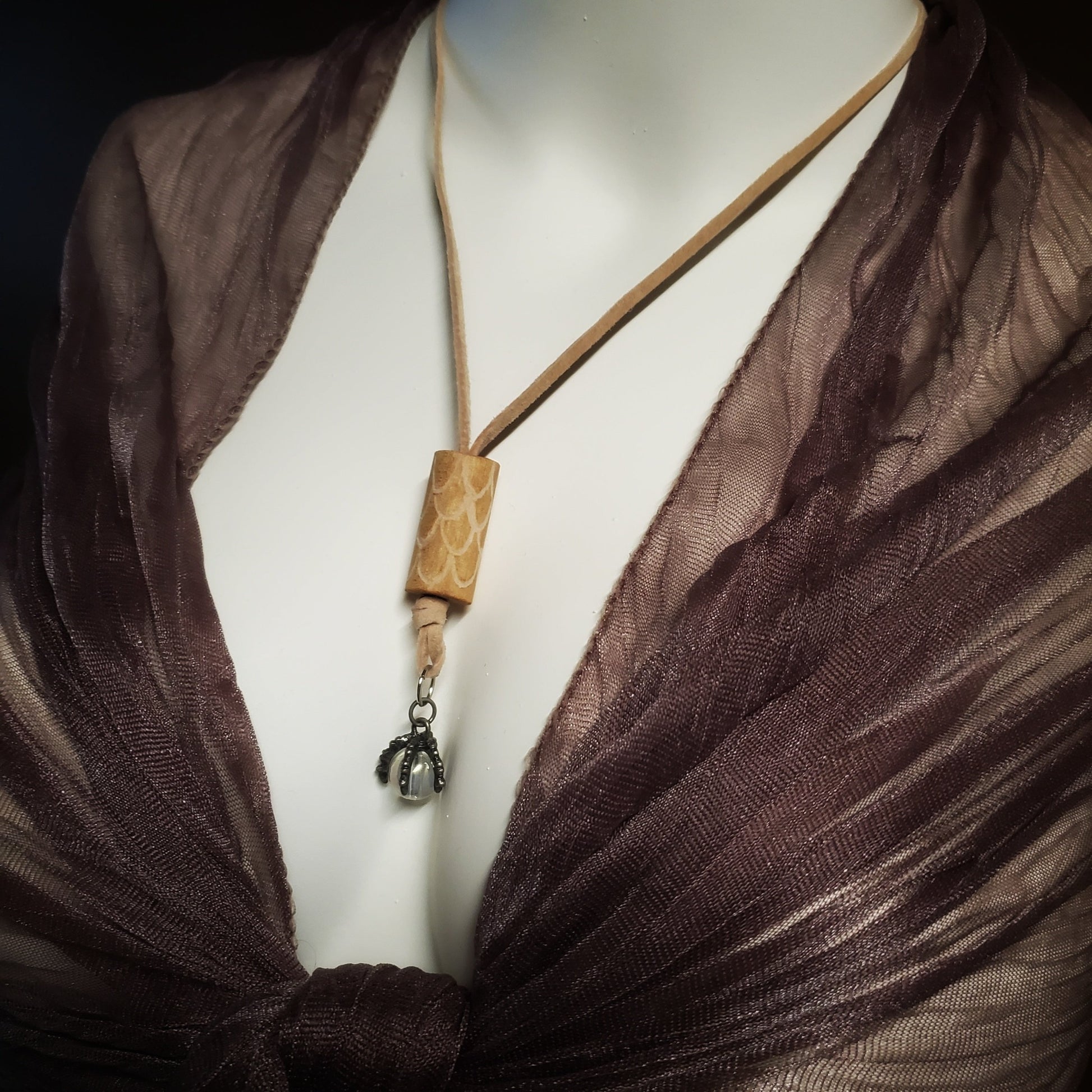 necklace made from a piece of upcycled drumstick on which dragon scales are etched under the drumstick a silver coloured dragon claw pendant holds a clear bead - a beige swede cord holds the pendant - the necklace hangs on the neck of a white mannequin bust wearing a sheer wrap