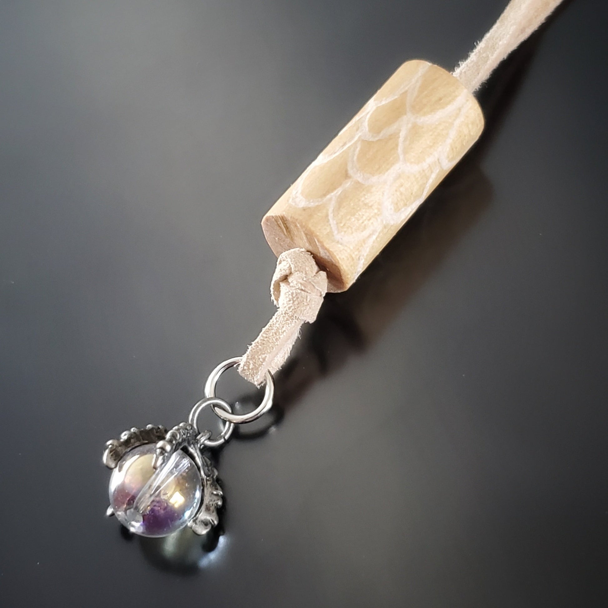 necklace made from a piece of upcycled drumstick on which dragon scales are etched under the drumstick a silver coloured dragon claw pendant holds a clear bead - a beige swede cord holds the pendant- black backgound