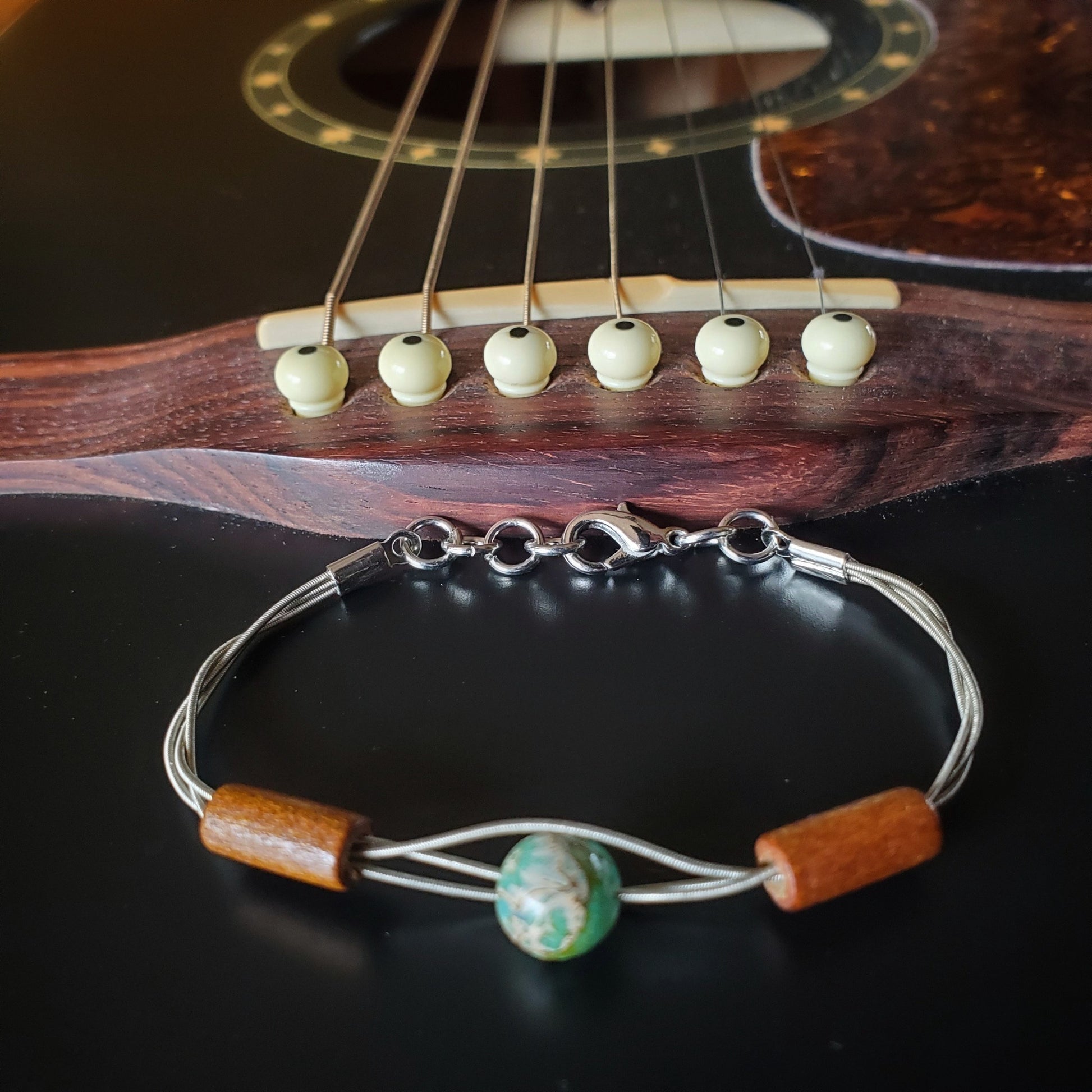 clasp style bracelet made from an upcycled guitar string on which there are two wood bead on either side of a green and gray round bead - the bracelet is in front of the bridge of a black guitar