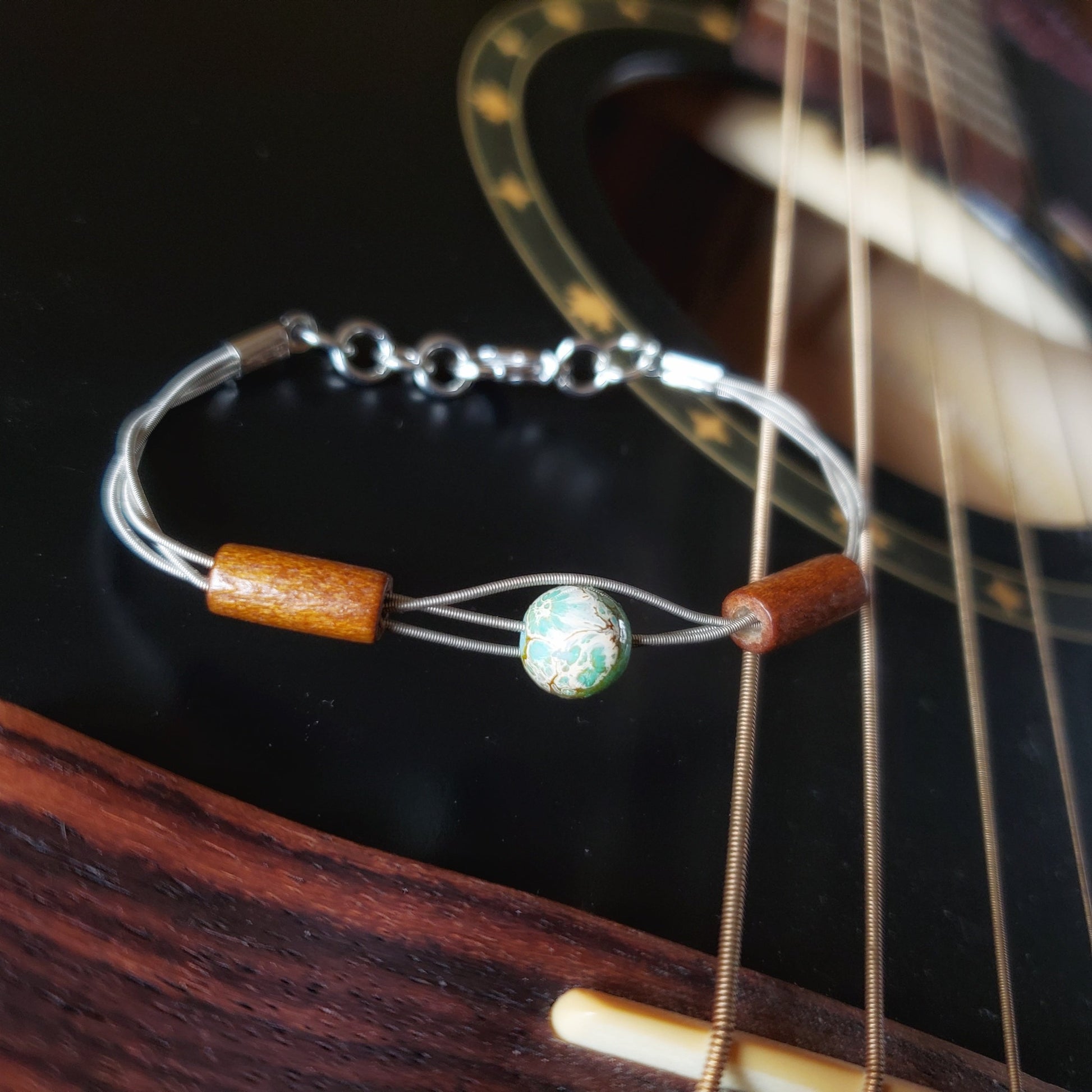 clasp style bracelet made from an upcycled guitar string on which there are two wood bead on either side of a green and gray round bead - the bracelet is resting on the string of a black guitar