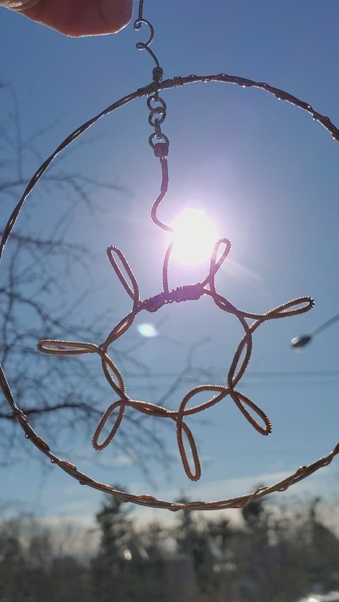 video of a a decoration shaped like a sun with a circle around it hanging off a chain - the sun and circle are made from upcycled guitar strings - sunshine and a blue sky are seen behind the decoration