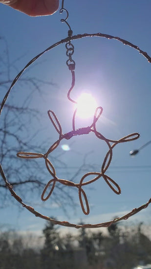 video of a a decoration shaped like a sun with a circle around it hanging off a chain - the sun and circle are made from upcycled guitar strings - sunshine and a blue sky are seen behind the decoration