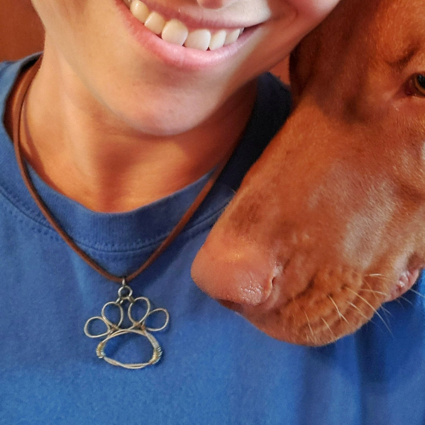 bottom of a woman's face wearing blue t-shirt and a necklace - necklace has a paw print pendant made from an upcycled guitar string - nose of a vizsla