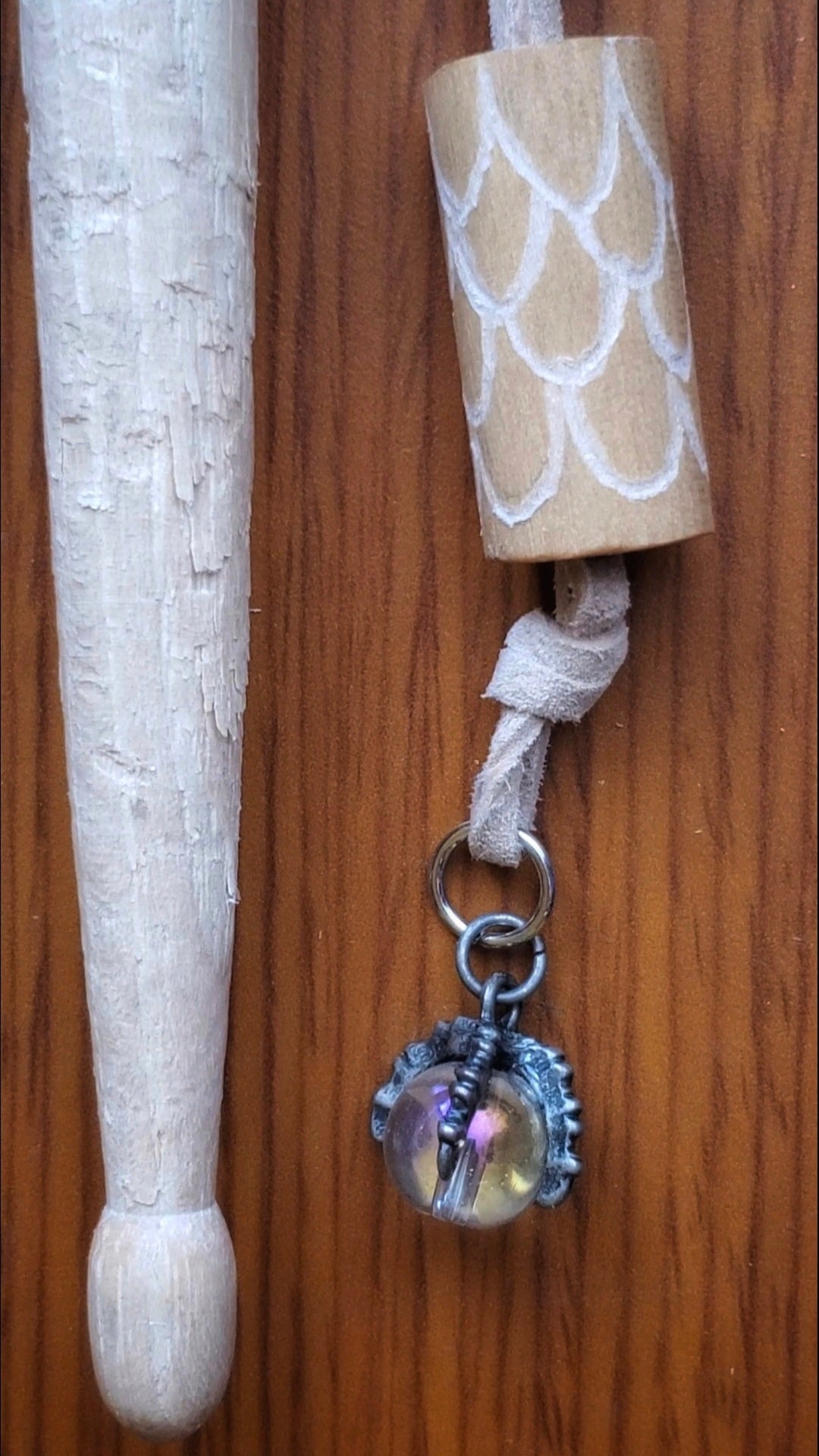 necklace made from a piece of upcycled drumstick on which dragon scales are etched under the drumstick a silver coloured dragon claw pendant holds a clear bead - a beige swede cord holds the pendant - next to the pendant there is a damaged drumstick