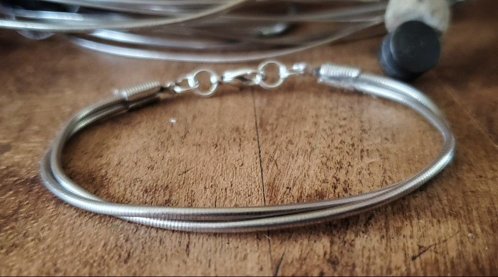 silver coloured clasp style bracelet made from upcycled upright bass strings - behind is a pile of upright bass strings