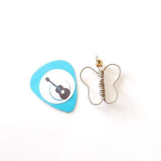 a pendant made from an upcycled guitar string shaped into a butterfly - the pendant sits next to a light blue guitar pick on which there is a white circle and the picture of a black guitar - white background