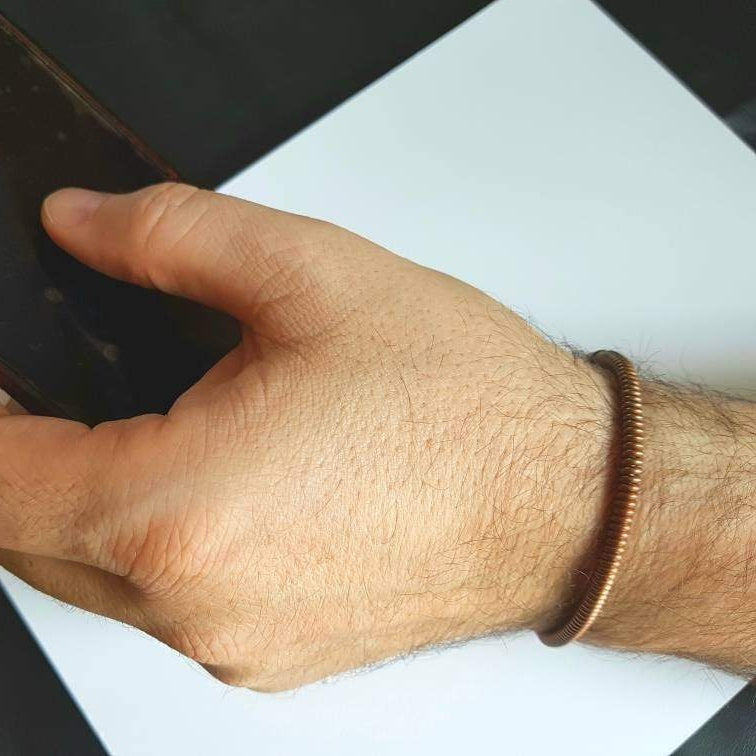 man's hand and wrist- he is wearing a copper cuff style bracelet made from an upcycled piano string and is holding a cell phone