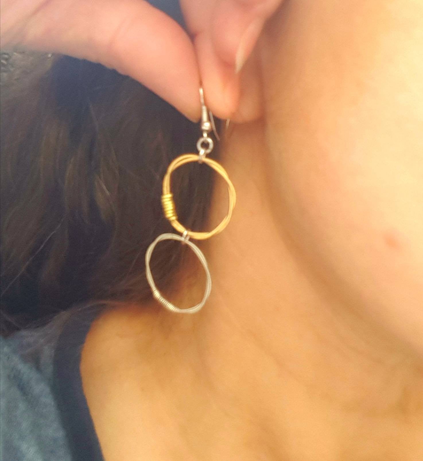 side of woman's head with brown hair, fingers are holding an earring near the ear, the top hoop of the earring is a gold coloured guitar string, the bottom hoop of the earring is made from a silver coloured guitar string