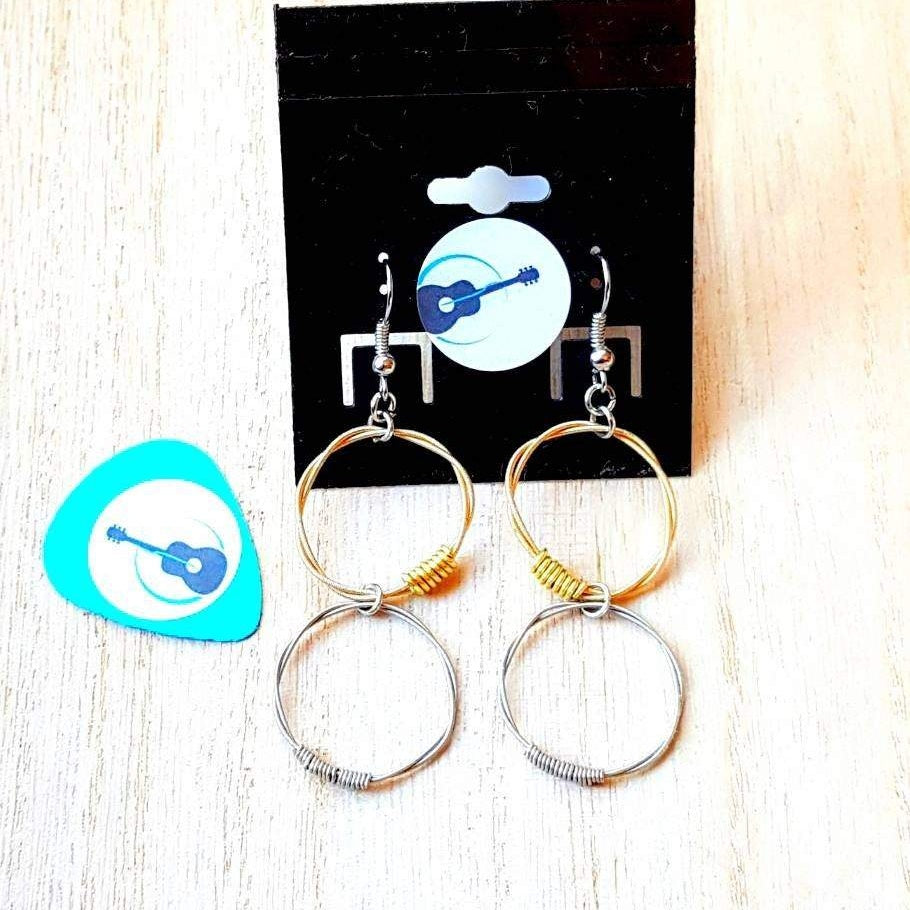 a pair of earrings with 2 hoops the top hoops are made from gold coloured guitar strings and the bottom ones are made from silver coloured guitar strings -they are hooked onto a black earring card- on the left is a blue guitar pick with an image of a black guitar