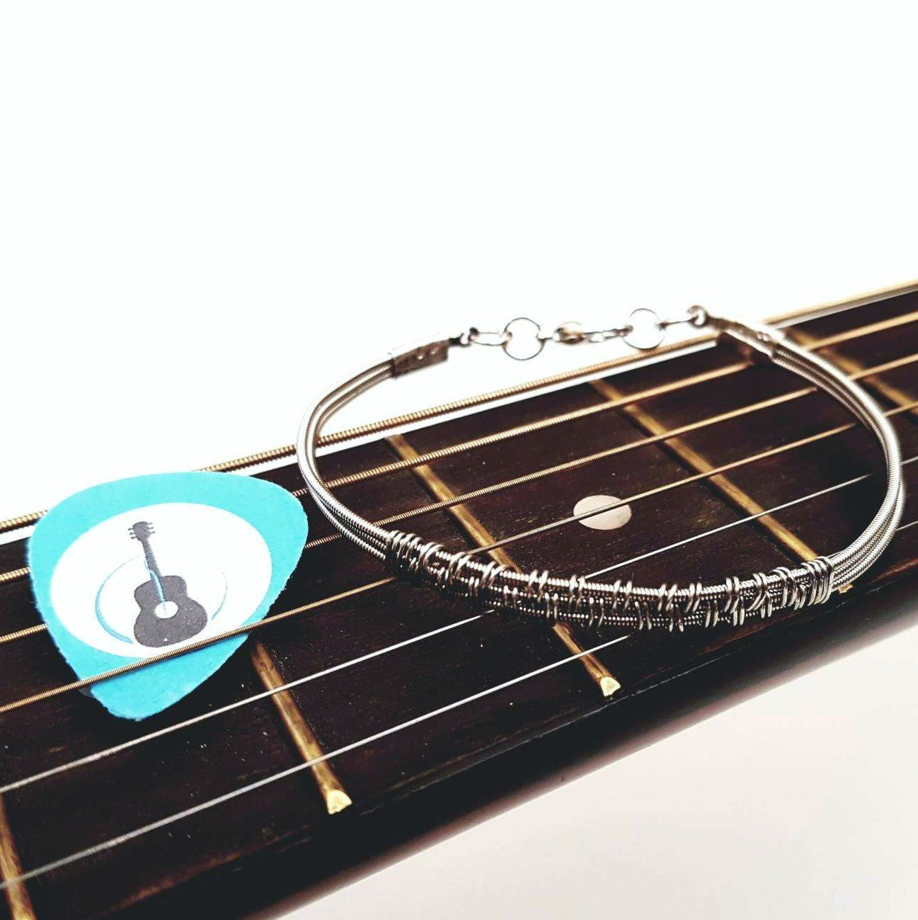 silver coloured clasp style bracelet made from 3 upcycled guitar strings - there is wire wrapped around the center of the bracelet - on the left is a blue guitar pick with an image of a black guitar- both are resting on the bridge of a guitar