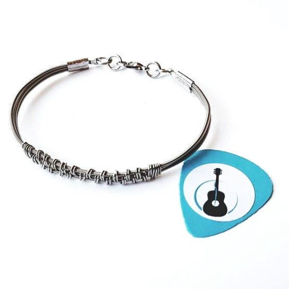 silver coloured clasp style bracelet made from 3 upcycled guitar strings - there is wire wrapped around the center of the bracelet in a pattern - next to it is a blue guitar pick with an image of a black guitar- white background