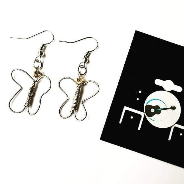 a pair of earrings made from upcycled guitar strings shaped into butterflies - the earrings are next to a black jewelry card which has a round white sticker with a picture of a black guitar and a blue circle - white background
