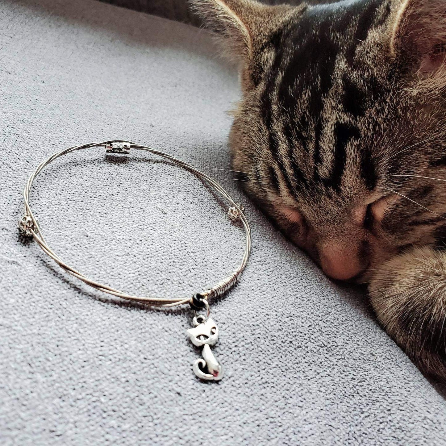 a silver coloured bangle style bracelet made from an upcycled guitar string on which a silver coloured cat shaped pendant hangs - a striped cat is sleeping next to the bracelet which sits on a grey background