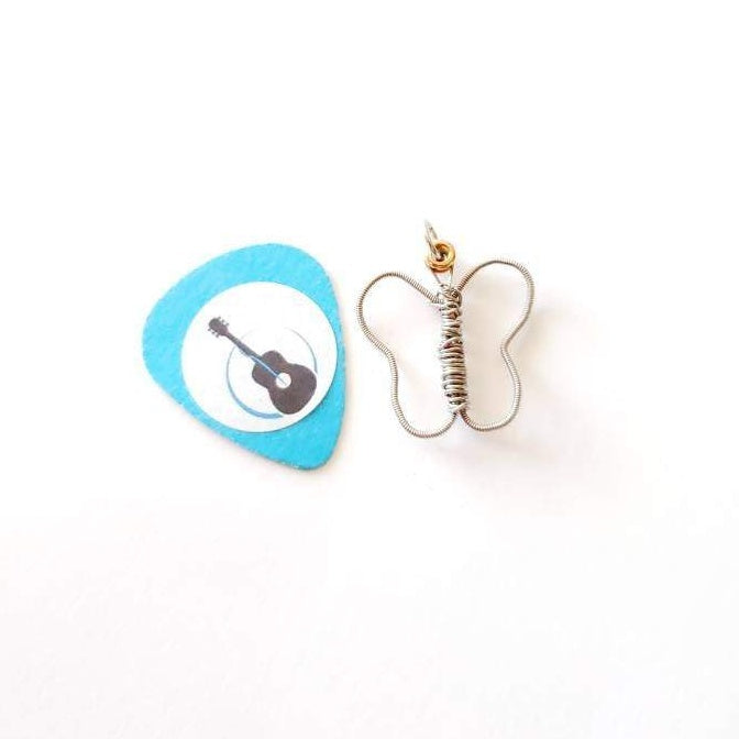a pendant made from an upcycled guitar string shaped into a butterfly - the pendant sits next to a light blue guitar pick on which there is a white circle and the picture of a black guitar - white background