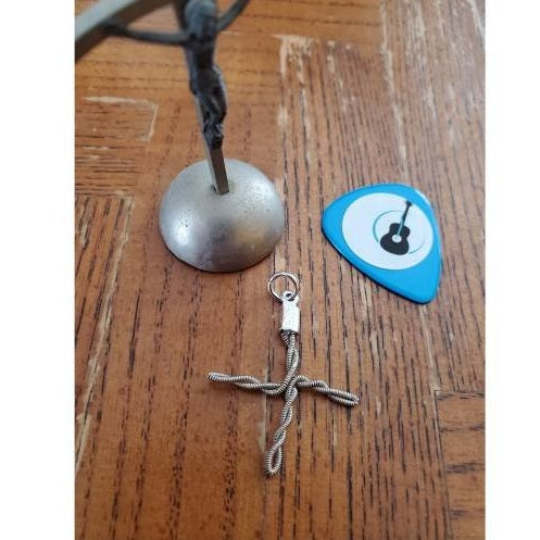 a cross shaped pendant made from an upcycled guitar string  -next to it is a blue guitar pick with a white circle and the picture of a black guitar - next to that is a silver coloured cross figurine