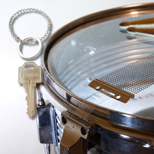 a keychain made from an upcycled snare drum string is above a silver key, on the right we see a snare drum on which we see part of a set of drumsticks and some snare drum strings - white background
