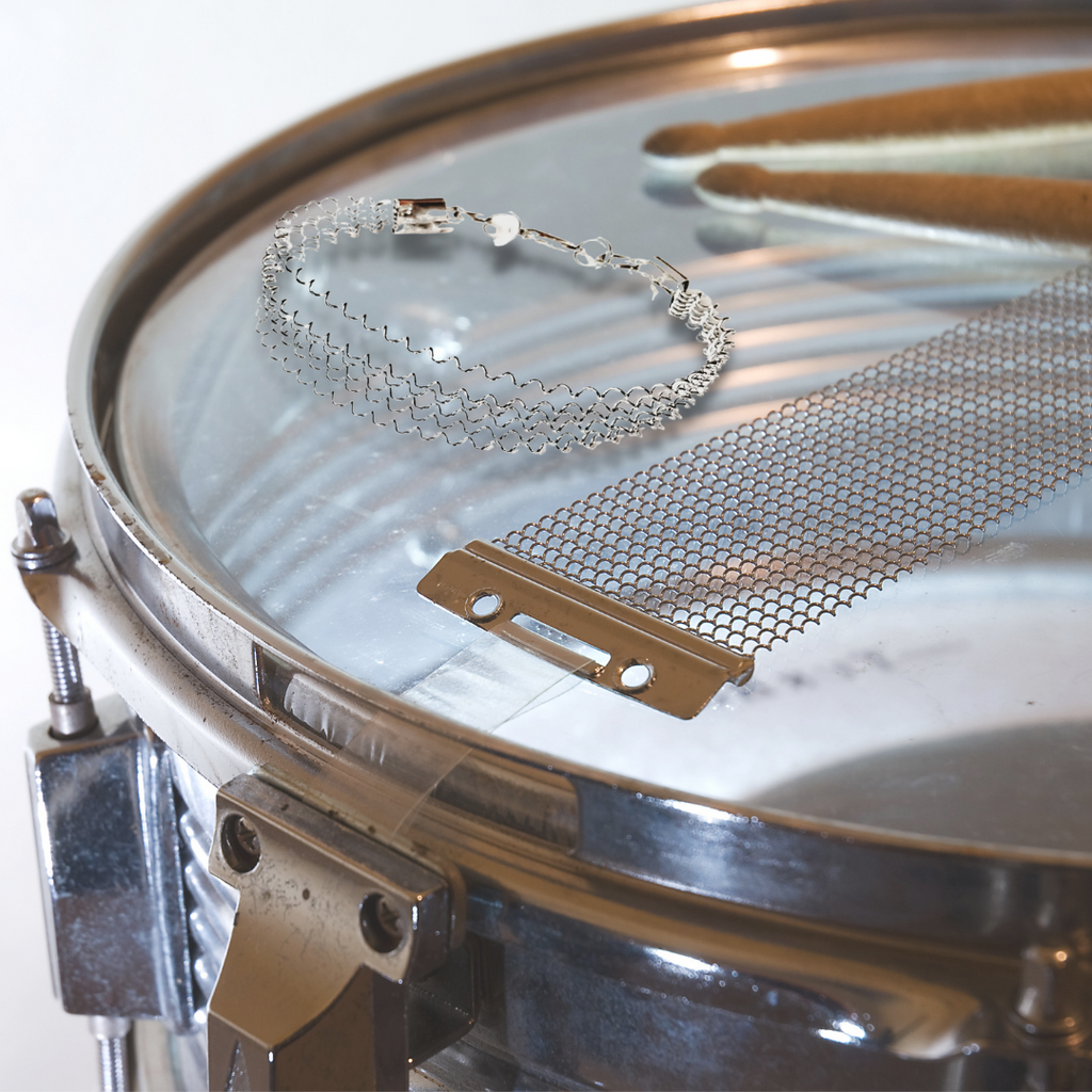 silver coloured clasp style bracelet made from a series of upcycled snare drum strings - bracelet is sitting on a snare drum which is also holding a set of drumsticks and a drum snare